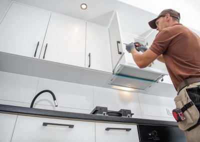 Man in a hat and gloves using a power tool to work on kitchen cabinets, with white cabinets, a black countertop, and polished marble flooring.