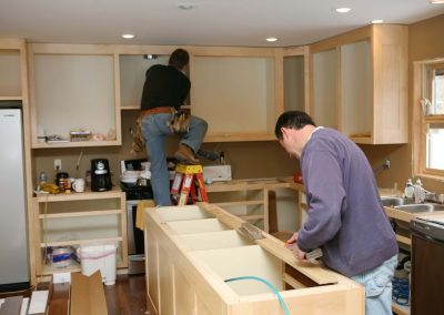 Two men are installing wooden kitchen cabinets in a kitchen under renovation, surrounded by tools and materials. A slab of granite is propped against the wall, ready to be fitted as the new countertop.