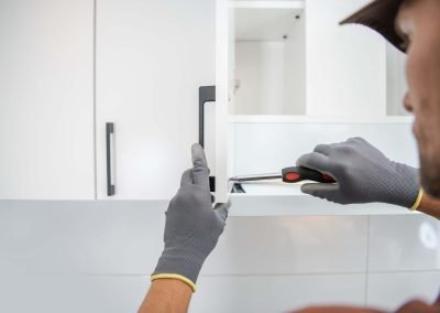 A person wearing gloves is using a screwdriver to assemble a white cabinet in a kitchen with marble flooring.