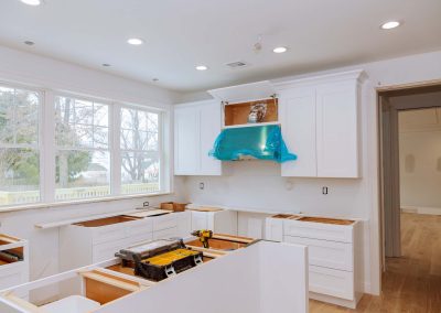 A kitchen under construction with white cabinets, a blue-covered range hood, and woodworking tools scattered. Gleaming marble countertops enhance the space, adding a touch of elegance to the soon-to-be-finished kitchen.