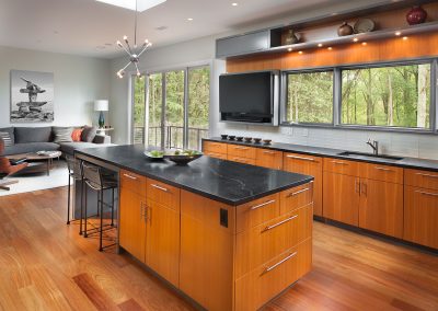 Modern kitchen with wooden cabinets, black countertops, an island with bar stools, and large windows offering a forest view. The elegant marble kitchen countertops complement the sleek flooring, making the space both stylish and functional.