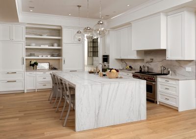 Modern kitchen with large marble island, pendant lights, white kitchen cabinets, built-in appliances, and light wood flooring.