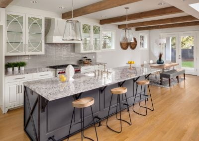 Modern kitchen with a large granite island, barstools, white kitchen cabinets, and wooden beams on the ceiling. Dining area in view.